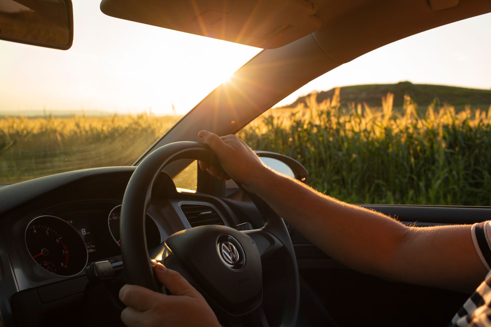 Bestuurder met de handen aan het stuur in een auto, langs een zonovergoten veld bij zonsondergang. Illustratieve foto.